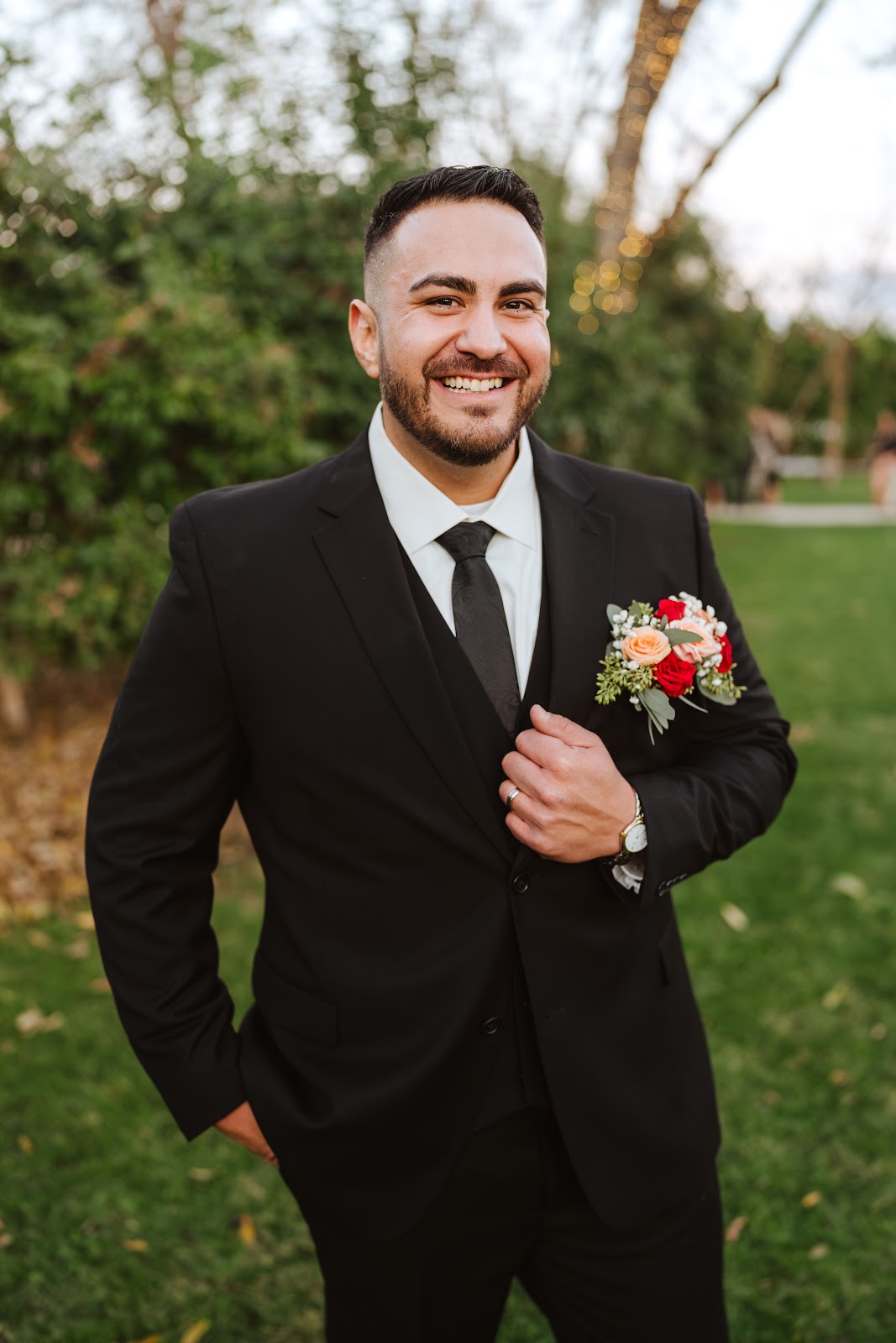 A groom in his black suit and boutonniere, smiling outdoors after his haircut